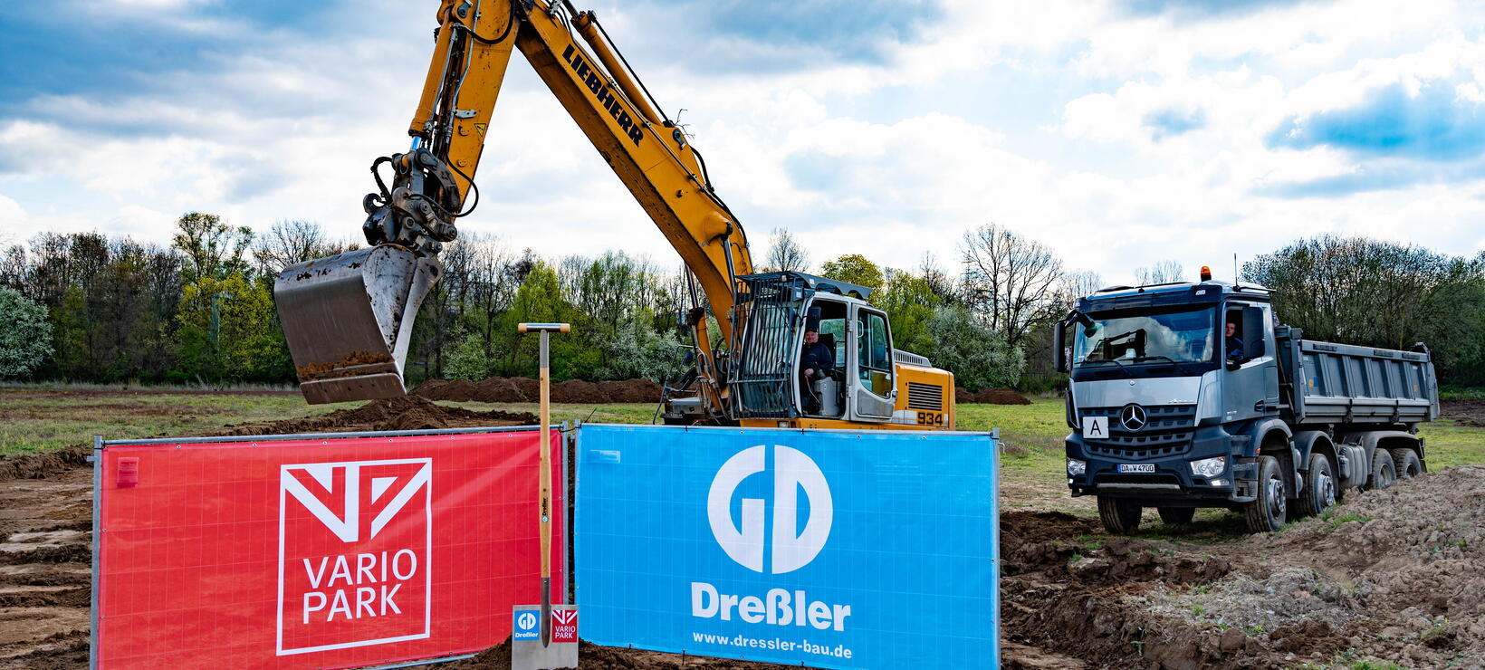 Ein riesiger Spaten mit den Logos der VarioPark Gmbh Und der Dreßler Bau GmbH in einem Erdhügel auf der Baustelle mit Bagger und Laster