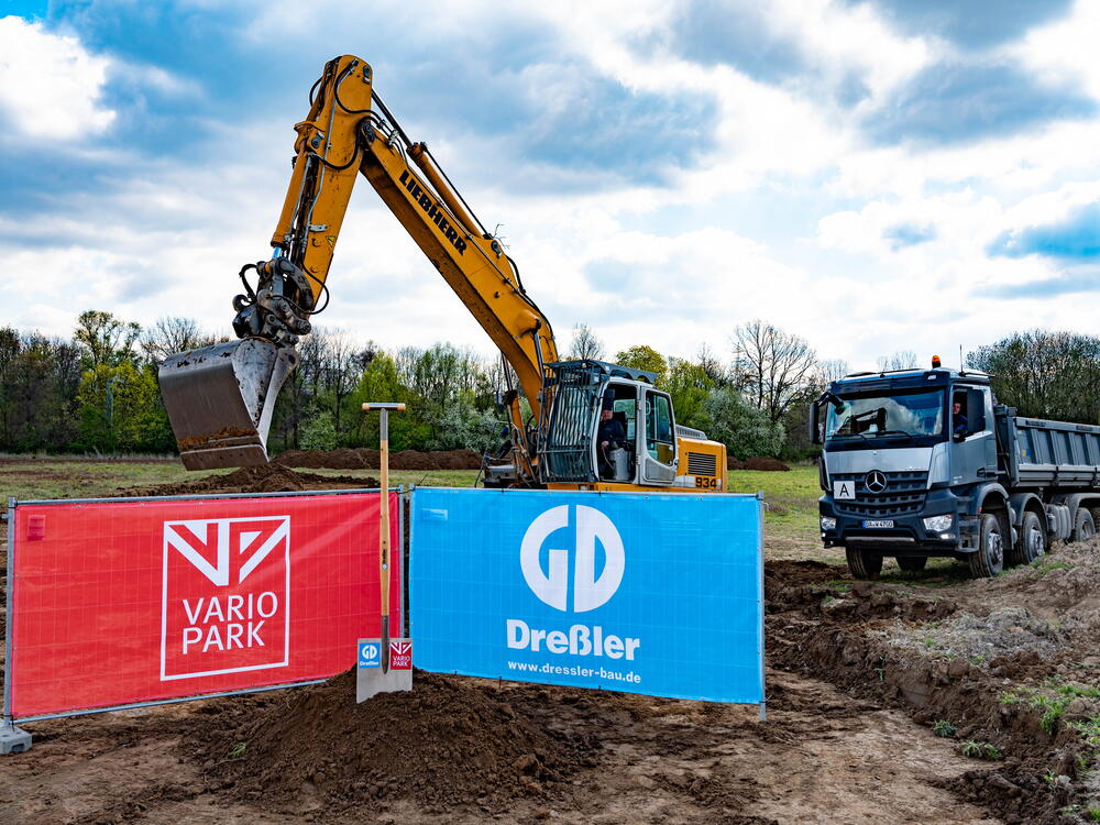 Ein riesiger Spaten mit den Logos der VarioPark Gmbh Und der Dreßler Bau GmbH in einem Erdhügel auf der Baustelle mit Bagger und Laster